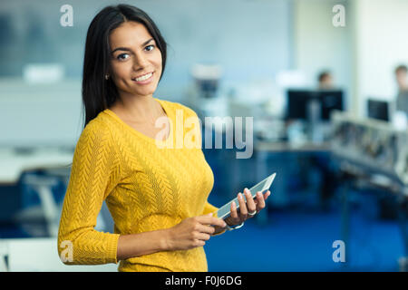 Ritratto di un allegro imprenditrice informale in un maglione in piedi con Tablet PC in ufficio e guardando la fotocamera Foto Stock