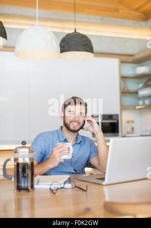 Uomo sorridente a bere caffè e parlando al cellulare al computer portatile Foto Stock