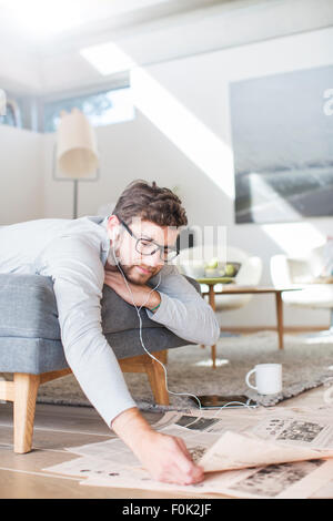 Uomo con cuffie leggere il giornale e bere il caffè nella stanza vivente Foto Stock