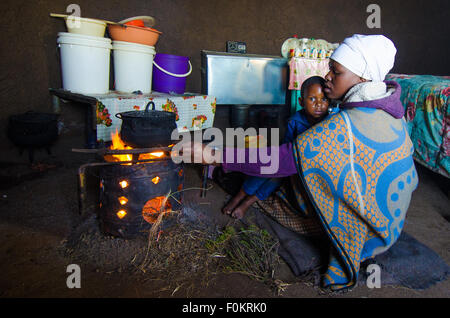 Un bambino curiosamente guarda come sua madre cucina un pasto su un piccolo una buca per il fuoco in una zona rurale Basotho home. Foto Stock