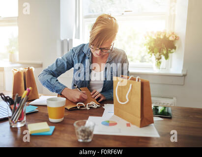 Giovane imprenditrice progettazione di imballaggi per il suo nuovo start-up business online seduto alla sua scrivania in casa a lavorare su una borsa regalo Foto Stock