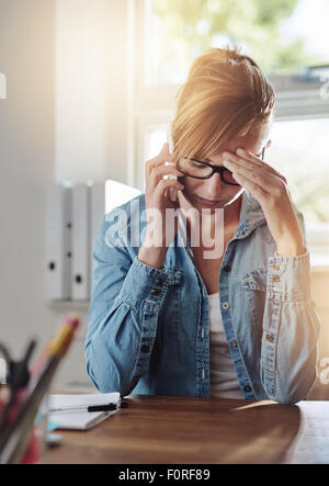 Interessato imprenditore femmina a parlare su un telefono come si siede alla sua scrivania nel suo ufficio a casa con la sua testa in appoggio sulla sua mano Foto Stock