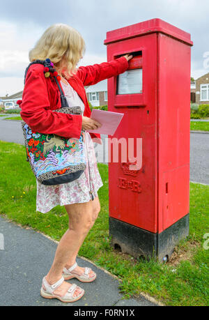 Donna di mezza età spedire una lettera in una forma rettangolare rossa letter box in Inghilterra, Regno Unito. Foto Stock