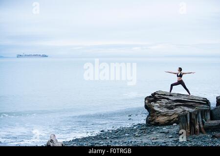 Donna matura la pratica dello yoga posa mentre permanente sulla grande driftwood ceppo di albero sulla spiaggia Foto Stock