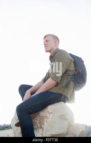 Giovane uomo che indossa uno zaino seduto sulla roccia che guarda lontano Foto Stock