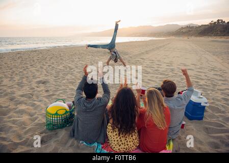 Gruppo di amici seduti sulla spiaggia, guardando amico do cartwheels su e, Tramonto, Vista posteriore Foto Stock