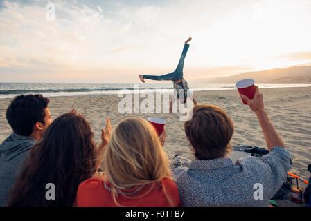 Gruppo di amici seduti sulla spiaggia, guardando amico do cartwheels su e, Tramonto, Vista posteriore Foto Stock