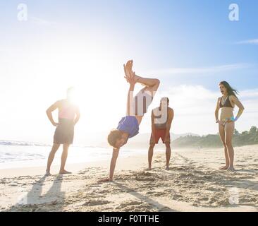 Gruppo di amici sulla spiaggia per guardare un amico fare handstand Foto Stock