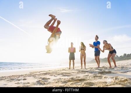 Gruppo di amici sulla spiaggia per guardare un amico fare somersault Foto Stock