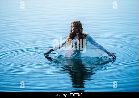 Giovane donna in piedi nel lago blu increspa la superficie con le dita Foto Stock