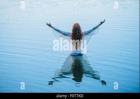 Vista posteriore della giovane donna in piedi nel lago blu con i bracci aperti Foto Stock