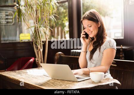 Metà donna adulta seduta utilizzando laptop e sorridente dello smartphone Foto Stock