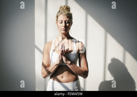 Una donna bionda, con gli occhi chiusi, in un bianco prodotto top e leggings, in piedi di fronte ad una parete bianca, fare yoga. Foto Stock