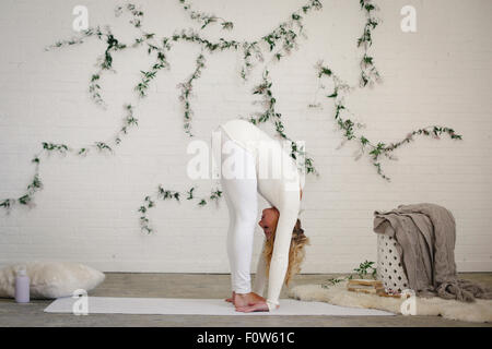 Una donna bionda, in un bianco leotard e gambali, in piedi su un tappetino bianco in una stanza in una posizione di yoga, la flessione in avanti e verso il basso. Foto Stock