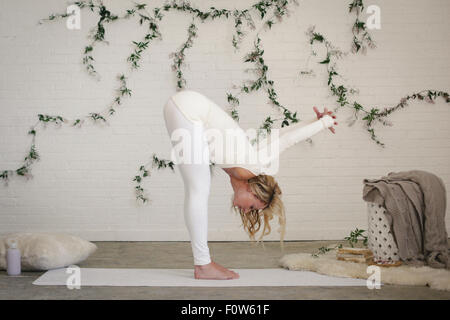 Una donna bionda in un bianco leotard e leggings la piegatura verso il basso allungamento. Un impianto di superriduttore sulla parete dietro di lei. Foto Stock