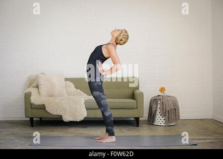 Una donna bionda in piedi su un materassino yoga in una stanza, fare yoga, flessione all'indietro con le sue mani sui fianchi. Foto Stock