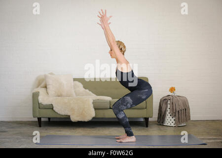 Una donna bionda in body e gambali, in piedi su un materassino yoga in una stanza, fare yoga, rannicchiato con le braccia sollevate. Foto Stock