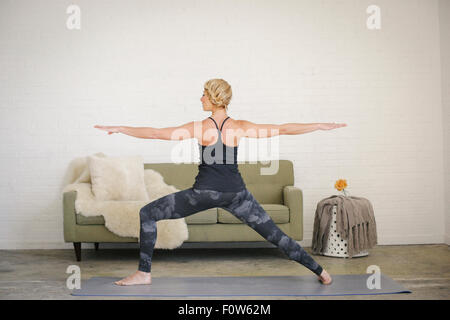 Una donna bionda in un nero leotard e gambali in piedi su un materassino yoga in una stanza, la sua gambe divaricate e braccia tese. Foto Stock