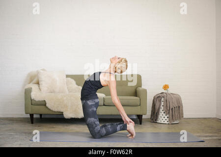 Una donna bionda inginocchiati su un materassino yoga in una stanza, fare yoga, flessione all'indietro con le sue mani toccano i suoi talloni. Foto Stock