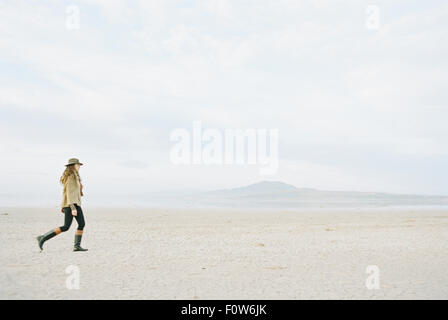 Donna con brown, lunga e capelli ricci a piedi attraverso una pianura desertica, indossando un cappello e stivali in pelle. Foto Stock