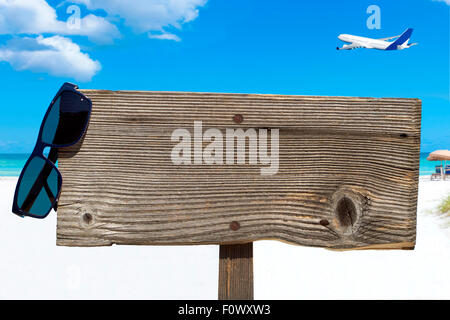 Cartello in legno con copia spazio e occhiali da sole sulla spiaggia soleggiata con un aereo nel cielo blu Foto Stock