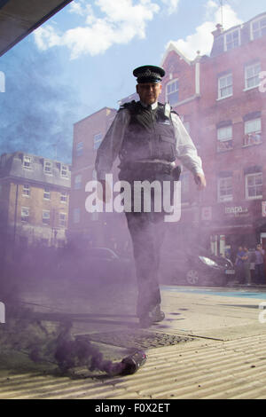 Cavo Street, Londra, 22 agosto 2015. Gli attivisti dalla guerra di classe dimostrare contro la neonata Jack lo Squartatore Museo sul cavo Street, affermando che denigrano le donne da off approfittando della morte di prostitute ucciso dall'aratro dissodatore. Il Museo, durante la sua fase di pianificazione, sostenuto che sarebbe dedicata al movimento Suffragettes. Nella foto: un funzionario di polizia calci una bomba fumogena lontano. Credito: Paolo Davey/Alamy Live News Foto Stock