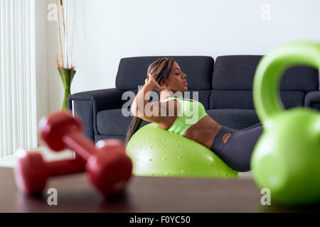 Giovane adulto americano africano donna in abbigliamento sportivo a casa, facendo fitness domestico e la formazione addominali su palla svizzera in l Foto Stock