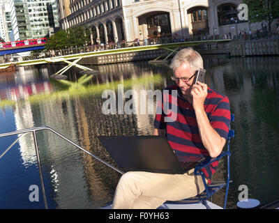 Uomo maturo al di fuori sulla sua barca parlando su iPhone 6 smartphone guardando al suo computer portatile Canary Wharf dietro a Londra E14 Foto Stock