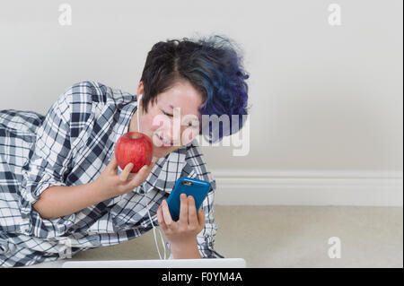 Teen ragazza con Apple mentre guarda il telefono cellulare con il computer in forefront distesi per ascoltare musica a casa. Foto Stock