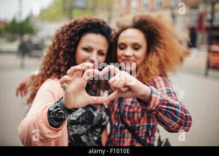 Felice giovani amici di sesso femminile messa a forma di cuore con le mani e le dita. Due donne in piedi insieme all'aperto sulla strada della citta'. La messa a fuoco Foto Stock