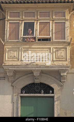Donna ad una finestra di un balcone in legno con ingresso al di sotto, il litorale di La Valletta, Malta. Foto Stock