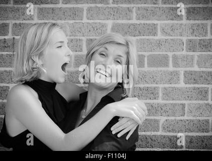 La figlia e la madre ridendo e scherzando insieme con i bracci intorno a ogni altro con un muro di mattoni in background Foto Stock