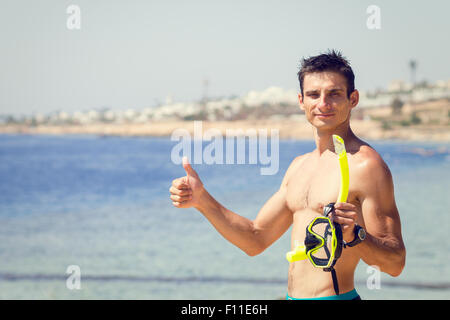 Giovane uomo con maschera e snorkel mostra pollice fino alla spiaggia del mare. Vacanze estive e snorkeling con uno sfondo con spazio di copia Foto Stock