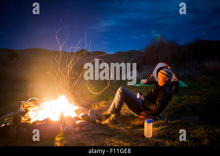 Uomo caucasico rilassante vicino al falò nel campo remoto, colline dipinte, Oregon, Stati Uniti Foto Stock