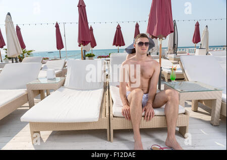 Giovane uomo che indossa gli occhiali da sole seduto sul bordo della sedia a sdraio sul ponte di lusso fronte Oceano Beach Resort Foto Stock