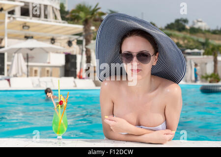 Giovane donna graziosa indossando occhiali da sole e cappello, appoggiata sul bordo di una piscina con bevande sul lato, e sorridente con la videocamera.. Foto Stock
