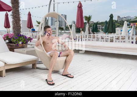 Per tutta la lunghezza della giovane uomo in costume da bagno effettuare una chiamata sul telefono cellulare mentre vi rilassate in vimini sedia a sdraio sul patio di fronte oceano di vacanza di lusso Resort. Foto Stock