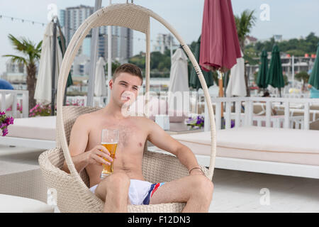 Ritratto di giovane uomo in costume da bagno rilassante nella sedia di vimini con bicchiere di birra fredda sul ponte di fronte Oceano vacanza di lusso Resort. Foto Stock