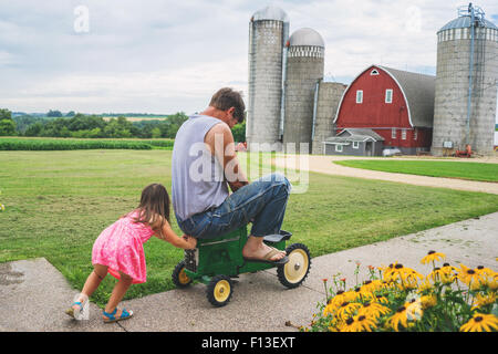 Ragazza che spinge suo padre su un trattore giocattolo in una fattoria, Stati Uniti Foto Stock