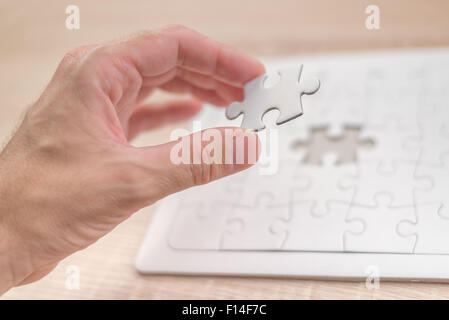 Maschio mettendo mano un pezzo mancante e risolvendo bianca puzzle collocato sulla sommità della vecchia quercia Legno tavolo, close up Foto Stock