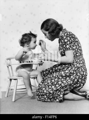 1930s inginocchiato madre Alimentazione bambino seduto in poltrona Foto Stock