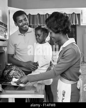 Anni sessanta famiglia americana africana IN CUCINA padre e figlia che guarda la madre staccare il tacchino arrosto dal forno Foto Stock