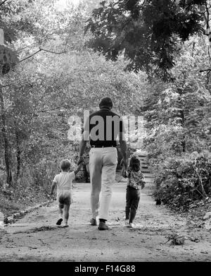 Anni sessanta vista posteriore padre tenendo le mani con due bambini il figlio e la figlia a piedi il sentiero del bosco Foto Stock