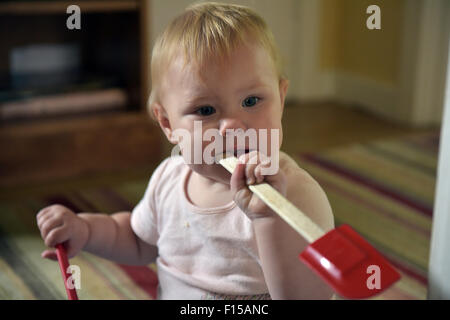 Un undici mesi di età bambino seduto sul pavimento di mordere su un cucchiaio (spatole) per alleviare il dolore e il disagio di mal di denti. Foto Stock