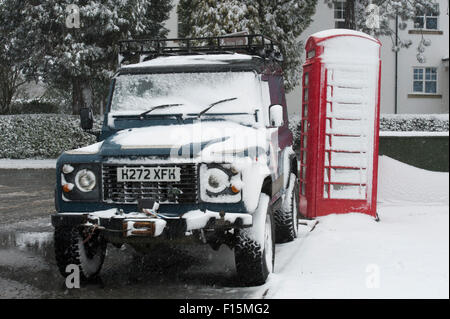 Due icone britannico coperto di neve bianca - Land Rover Defender 90 parcheggiato a fianco di un rosso inglese casella telefono su un inverno nevoso giorno - Ilkley, Inghilterra, Regno Unito. Foto Stock