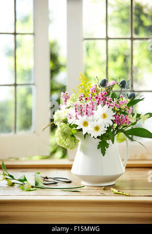 Bouquet di fiori recisi freschi contenenti Daisy, Flox, Mare Holly, ortensie, e l ambrosia in bianco brocca vaso sul davanzale della finestra Foto Stock