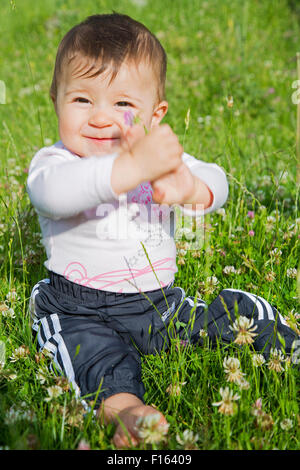 Un bambino di sei mesi ragazza seduta su un prato e mostrando un fiore nelle sue mani Foto Stock