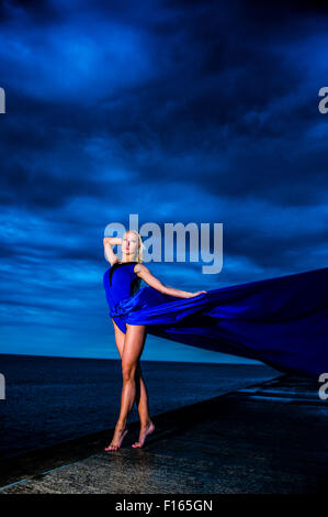 Posizione esterna fotografia - un alto slim bionda bionda donna modello ragazza in posa all'aperto su una serata umida con un blu lungo abito fluente fluttuante nel vento Foto Stock