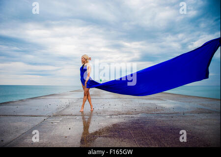 Posizione esterna fotografia - un alto slim bionda bionda donna modello ragazza in posa all'aperto su una serata umida con un blu lungo abito fluente fluttuante nel vento Foto Stock