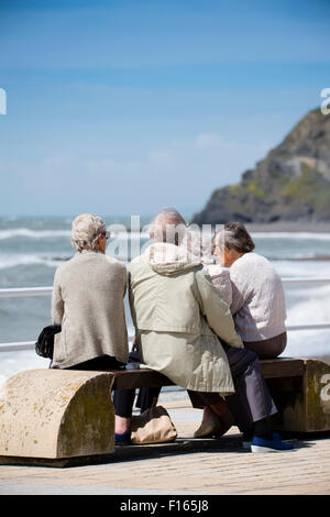 Un gruppo di quattro capelli grigi senior donne anziane amici seduti insieme su un banco di mare su un blustery pomeriggio estivo, indossando simile grigio taupe vestiti colorati, Aberystwyth Wales UK Foto Stock